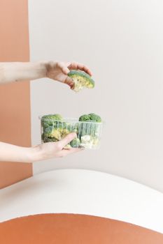 Close-up of hands holding broccoli in a plastic container against a minimalist backdrop.