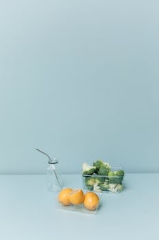 A fresh display of broccoli and oranges with a glass bottle on a light blue background.