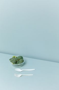 A minimalist image of fresh broccoli in a bowl with plastic cutlery, isolated on a light pastel background.