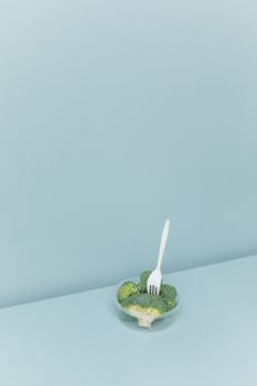 一个透明的碗里装着西兰花，旁边放着一把叉子，摆放在蓝色表面上，照片采用高角度简约风格拍摄