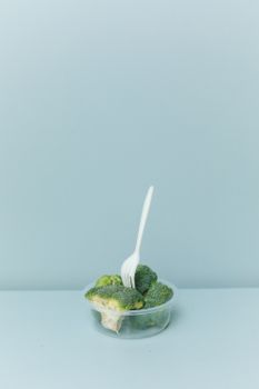 Fresh broccoli in a plastic container with a fork against a blue background.