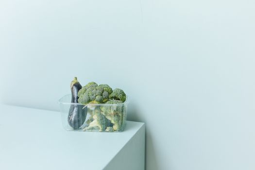 A plastic container with organic broccoli and eggplant on a minimalist white surface.