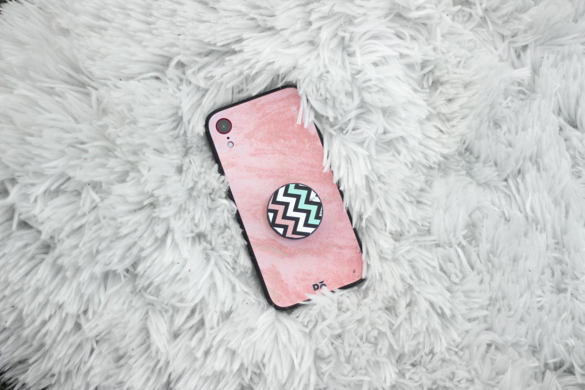 Top view of a mobile phone with a pink case and zigzag pop socket on a fluffy white rug.