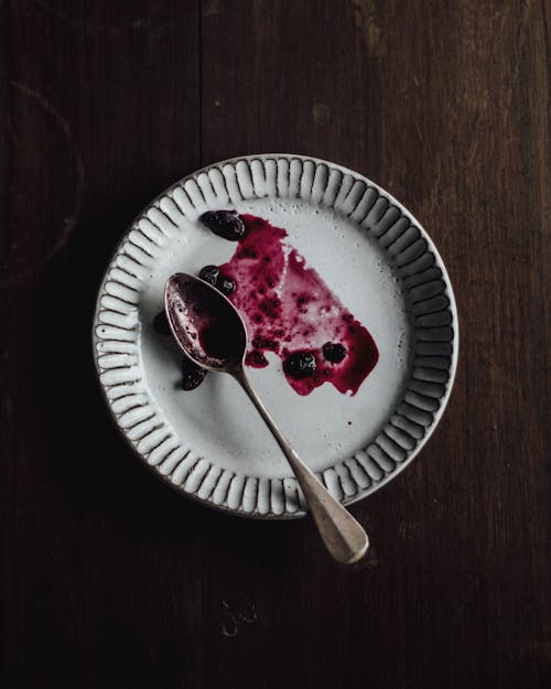 Top view of white empty plate with spoon and spread leftover blueberry sauce placed on wooden table in kitchen in morning