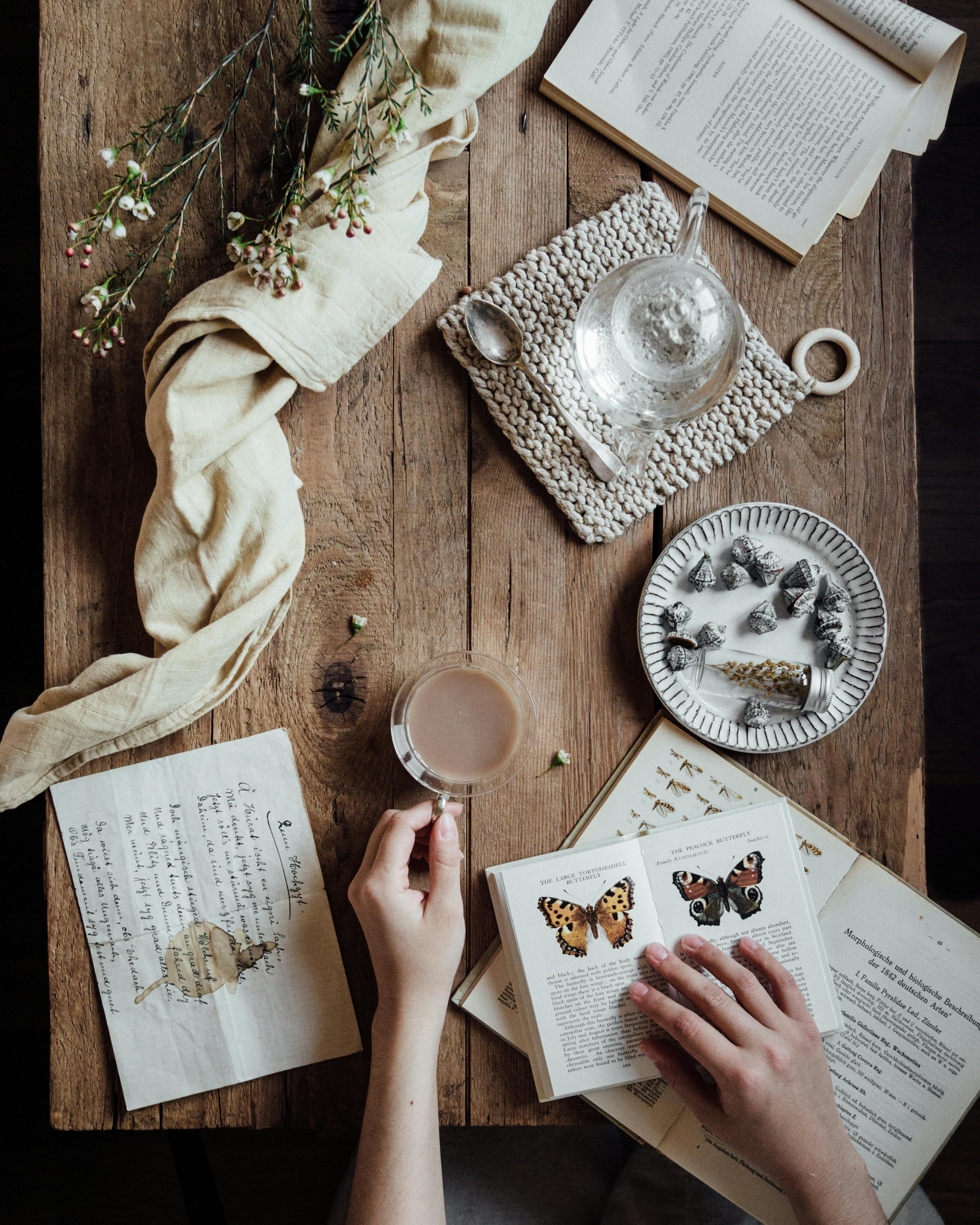A serene scene showing a book, tea, flowers, and hands on a wooden table.