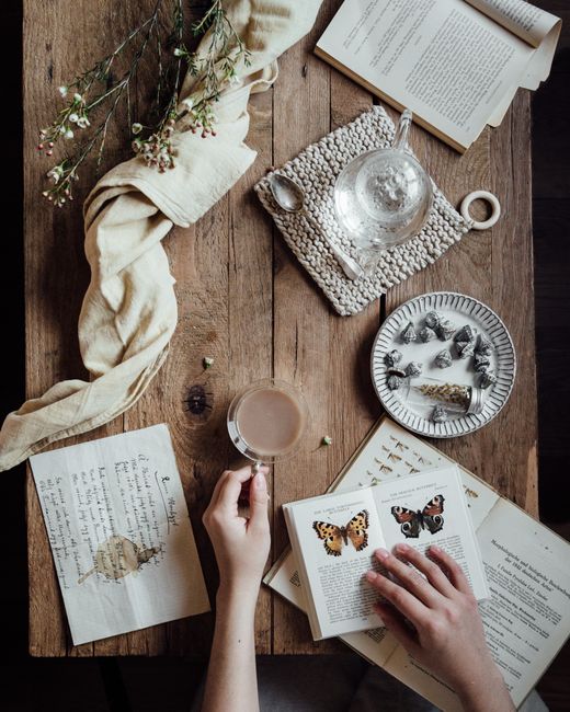 A serene scene showing a book, tea, flowers, and hands on a wooden table.