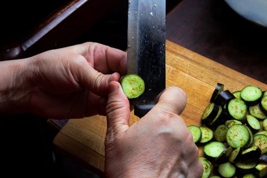 Hands slicing fresh eggplant on a wooden chopping board in natural light.