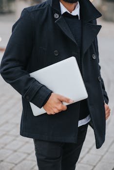 Stylish man in dark coat holding a laptop on a city street, representing modern professional lifestyle.