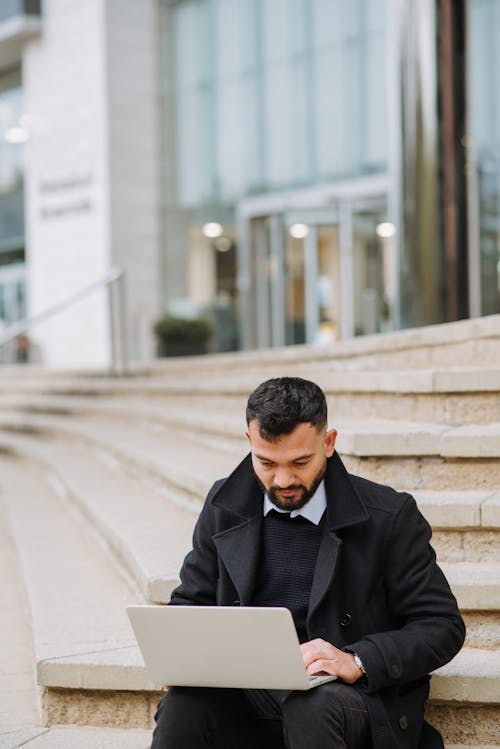 Free Young serious unshaven ethnic male manager in formal wear working on netbook on city staircase Stock Photo