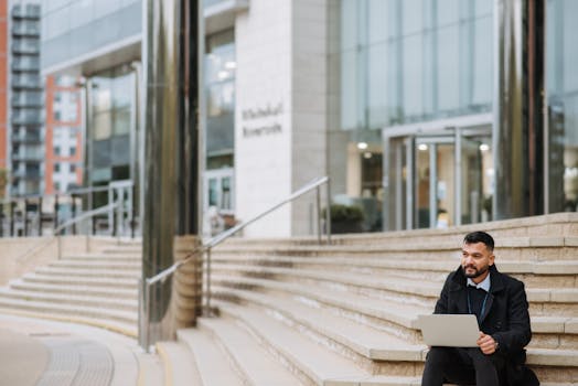 Thoughtful bearded ethnic male office worker with portable computer looking away on urban stairs against modern building