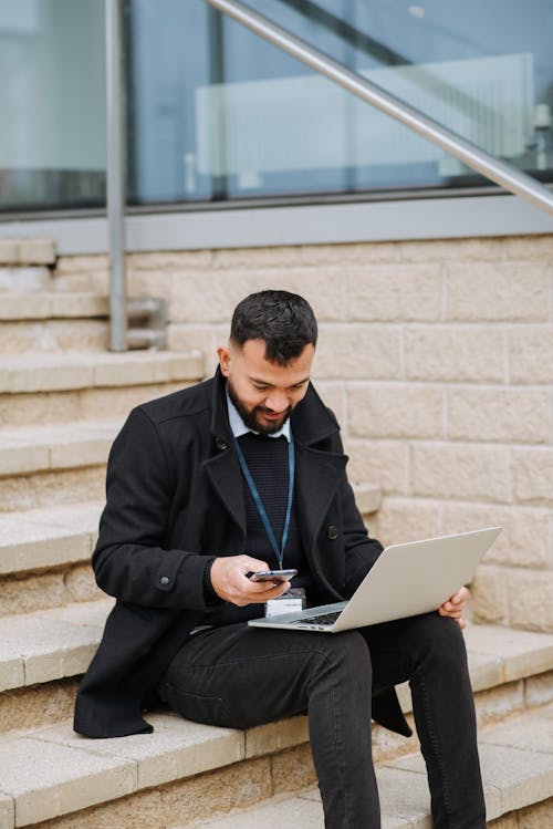 Attentive ethnic manager with laptop chatting on smartphone on staircase