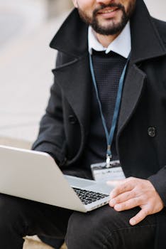 Adult male with beard using a laptop outside wearing a black coat and lanyard, representing modern work culture.
