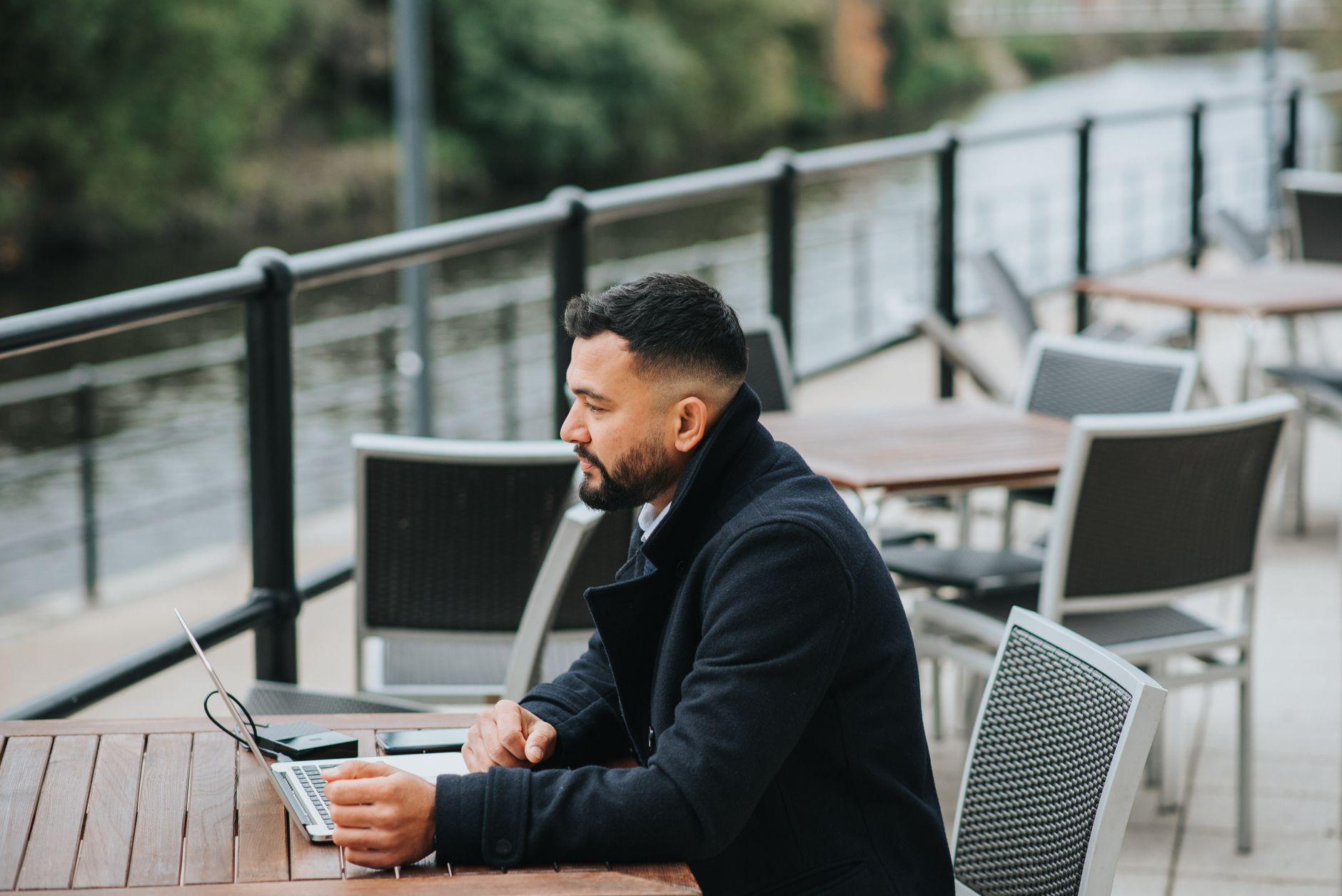Side view of young masculine pondering ethnic male entrepreneur sitting at table with netbook and looking forward on veranda of cafeteria