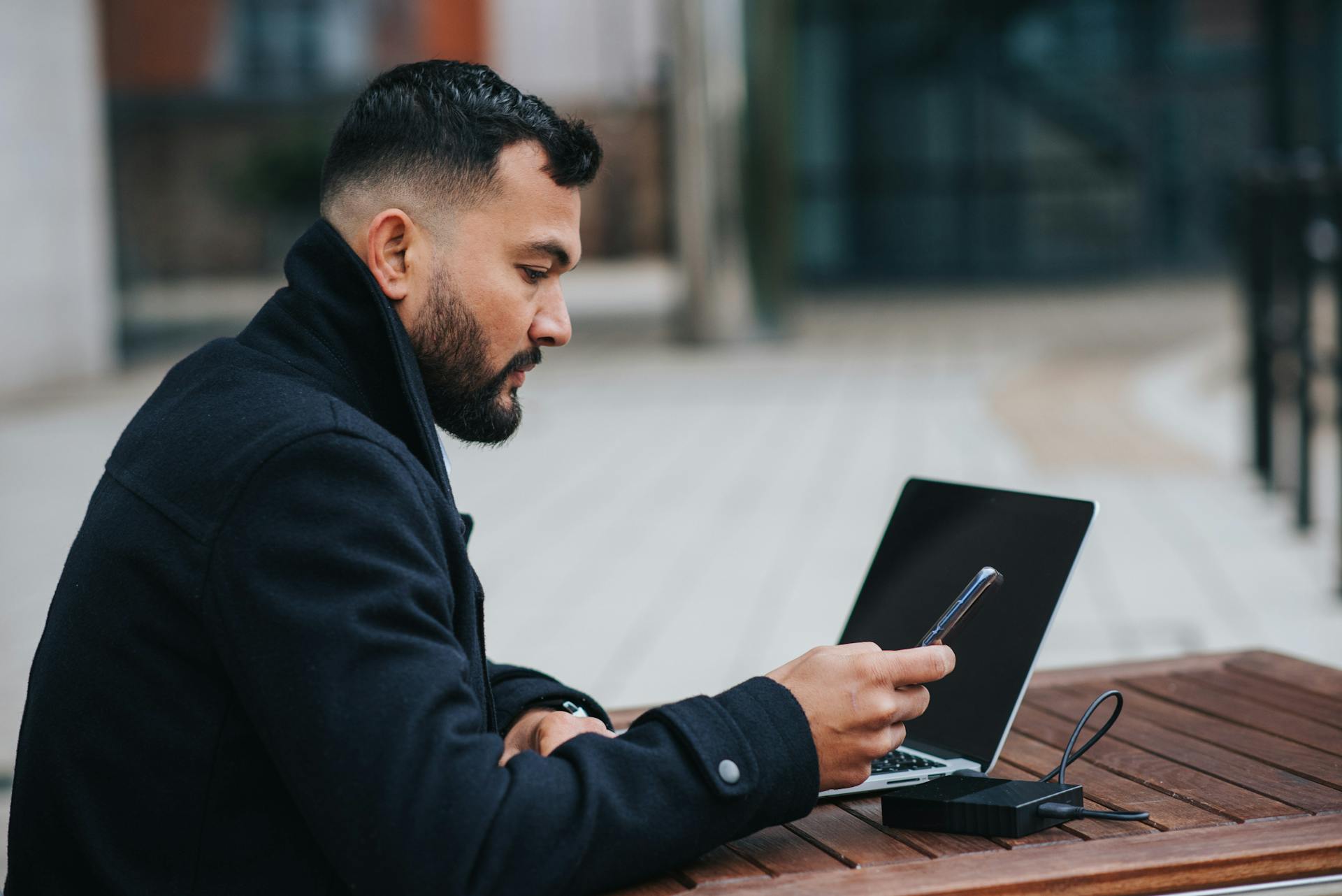 Side view of young concentrated ethnic male manager surfing internet on cellphone at cafe table with netbook and power bank on street