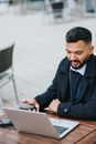 Young attentive ethnic male manager with cellphone watching netbook at cafe table in town