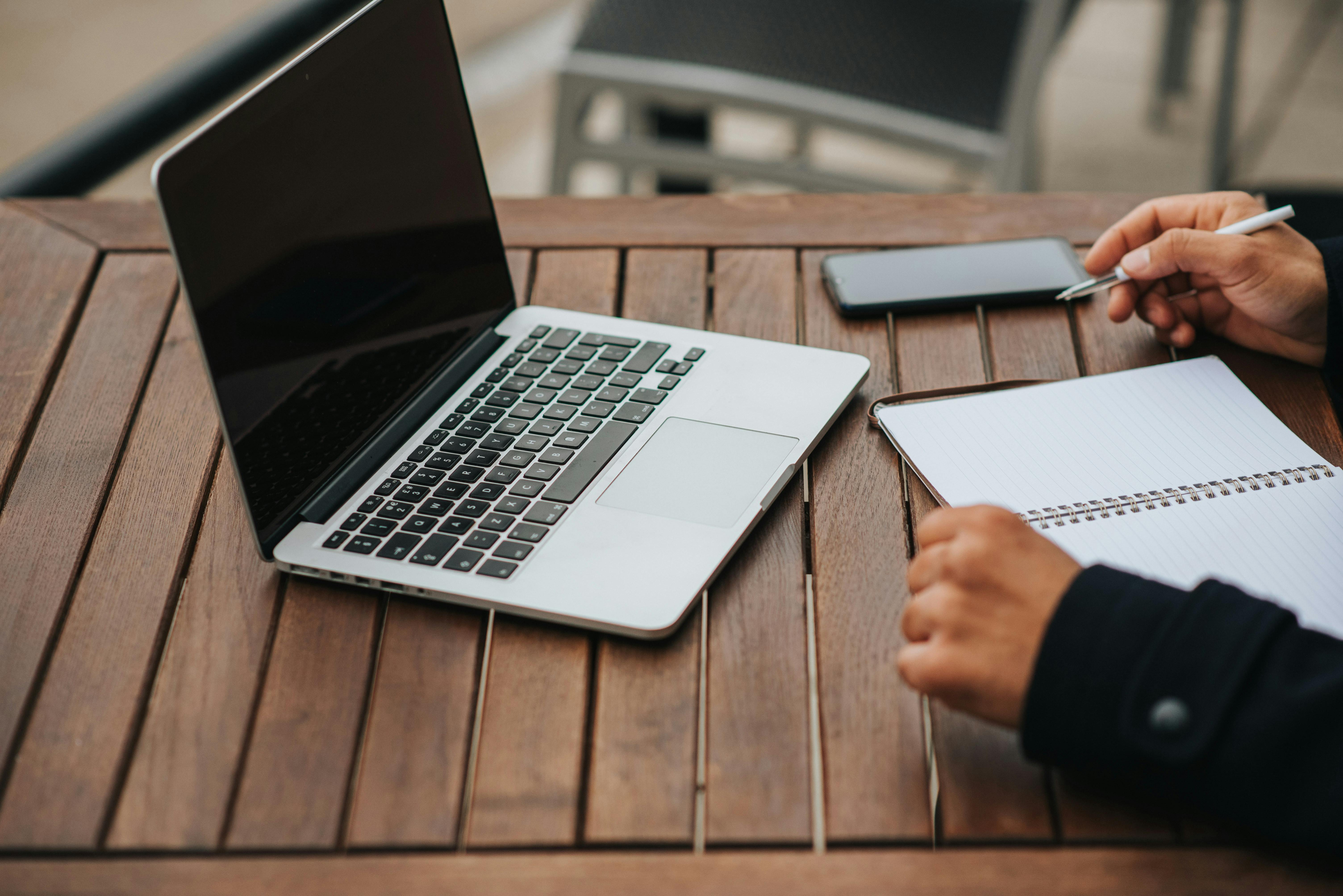 Open laptop and a person writing in a notebook on a wooden table, ready for remote work.