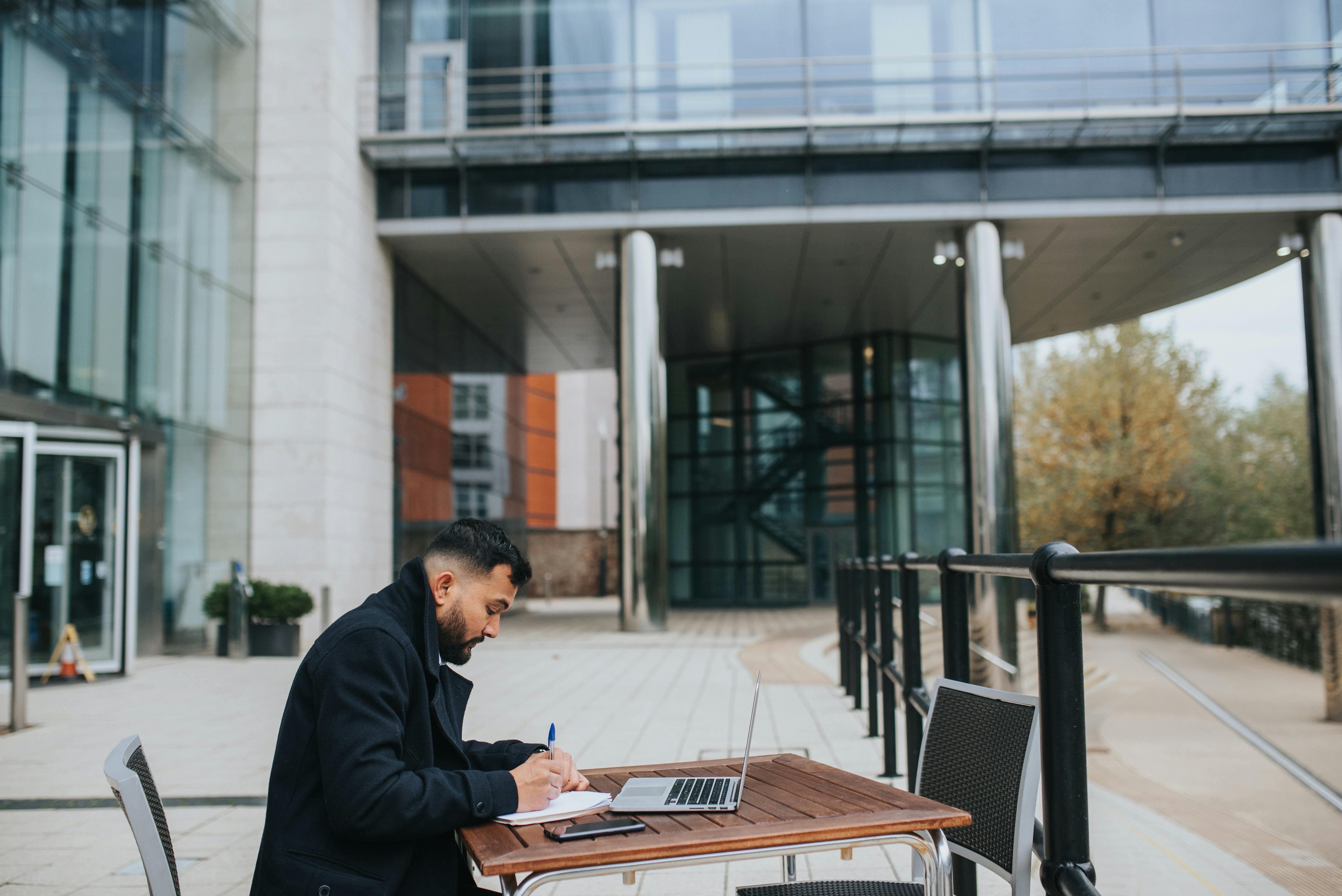 ethnic distance employee writing in copybook near laptop at table