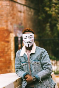 一名无法辨认的黑人男性穿着休闲装，戴着装饰面具，在城市中靠在栅栏上