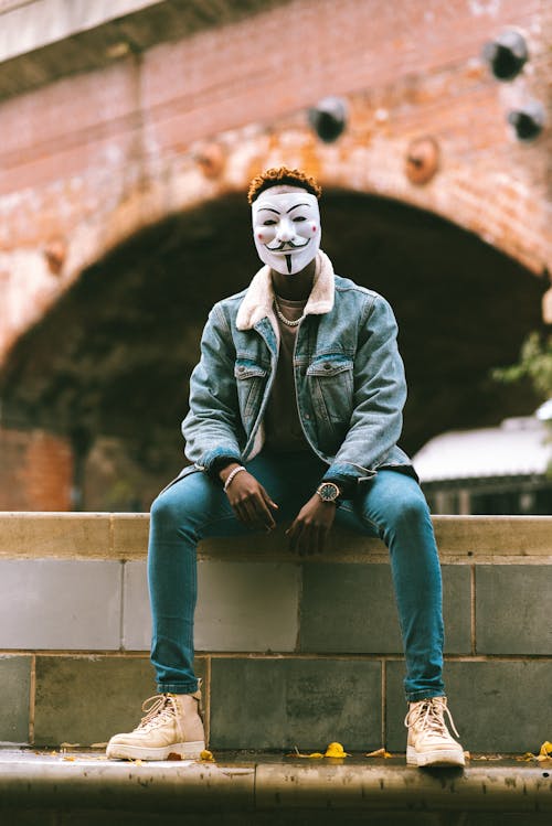 Free African American male in Anonymous mask and stylish clothes sitting on stone fence on street against brick construction in daytime Stock Photo