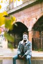 Unrecognizable black activist in Anonymous mask and stylish clothes sitting on stone fence on street and looking at camera