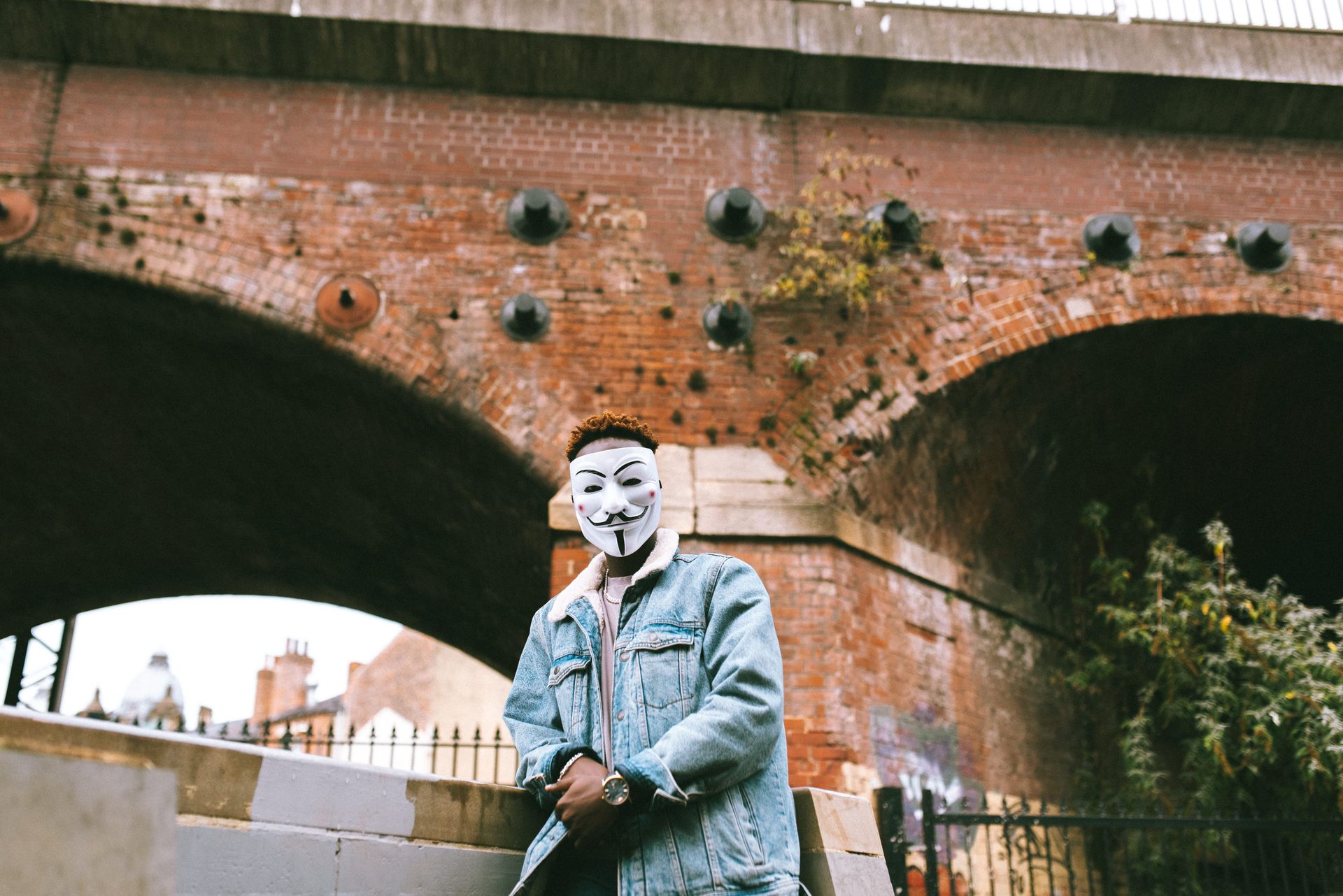 Man with Anonymous mask standing under brick archway in urban setting, symbolizing protest and anonymity.