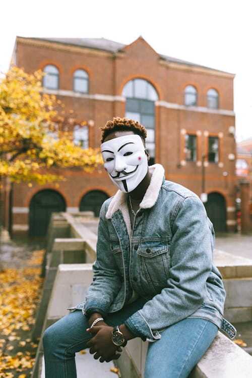 Free Concerned African American man in famous Anonymous mask sitting on stone fence against aged brick building and tree with bright autumn tree in daytime Stock Photo