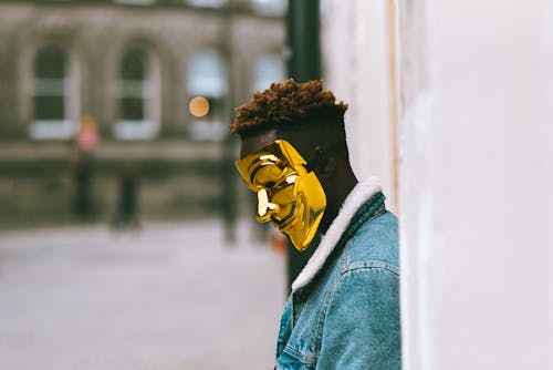 Free Side view of thoughtful African American activist wearing golden Anonymous mask as symbol of protest against current state policy standing near building on city street in daytime Stock Photo