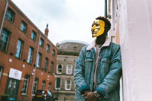 Free From below of confident black activist in stylish outfit and famous Anonymous mask leaning on building placed in city district in daytime Stock Photo
