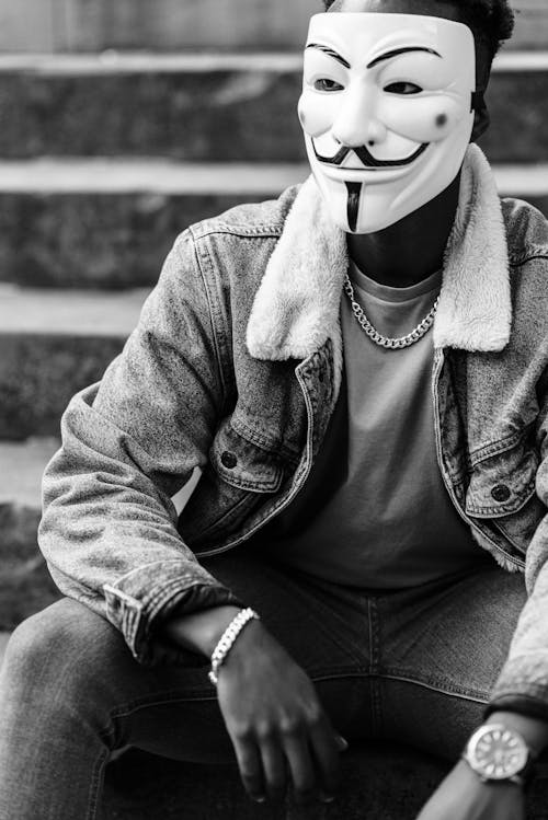 Free Black and white of crop black activist in famous Anonymous mask and stylish clothes sitting on stone stairs and looking away Stock Photo