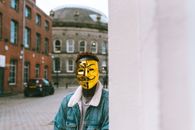 Unrecognizable African American activist in golden Anonymous mask standing near building on street in town and looking at camera