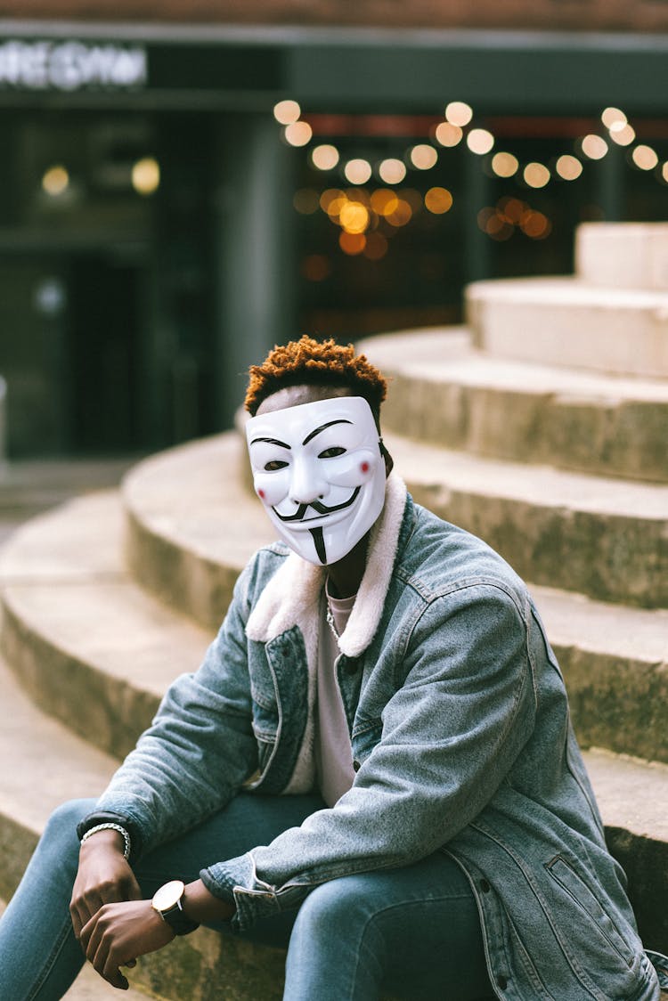 African American Guy In Anonymous Mask Sitting On Stairs On Street