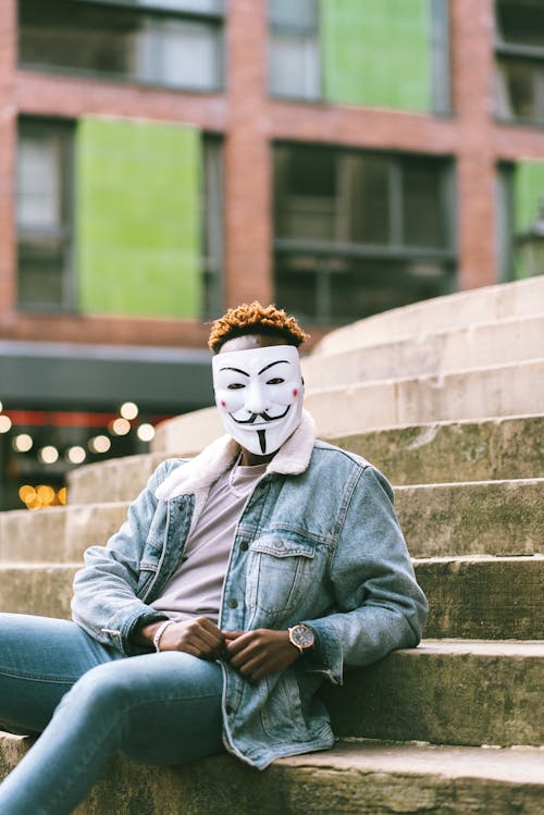 Free Unrecognizable African American activist in white Anonymous mask and stylish denim clothing sitting on stone stairs on street in city and looking at camera Stock Photo