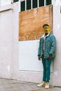Full body of African American guy in stylish denim clothes and golden Anonymous mask standing alone on street near building in daytime