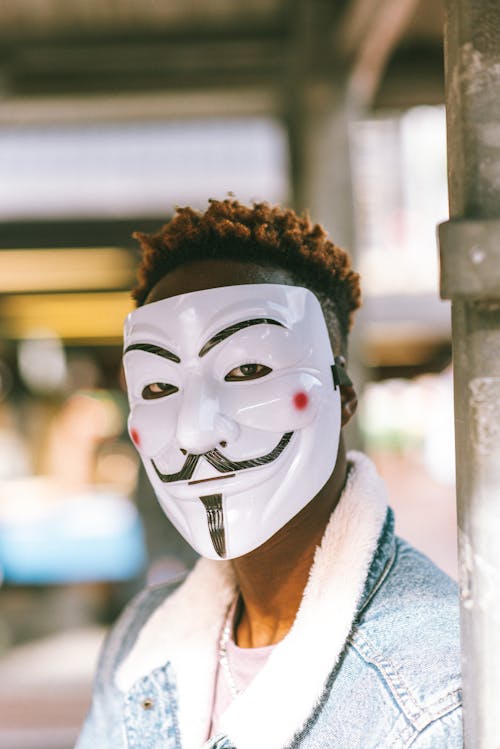 Free Worried black male wearing white Anonymous mask as symbol of protest against current state policy and standing on street in daytime Stock Photo