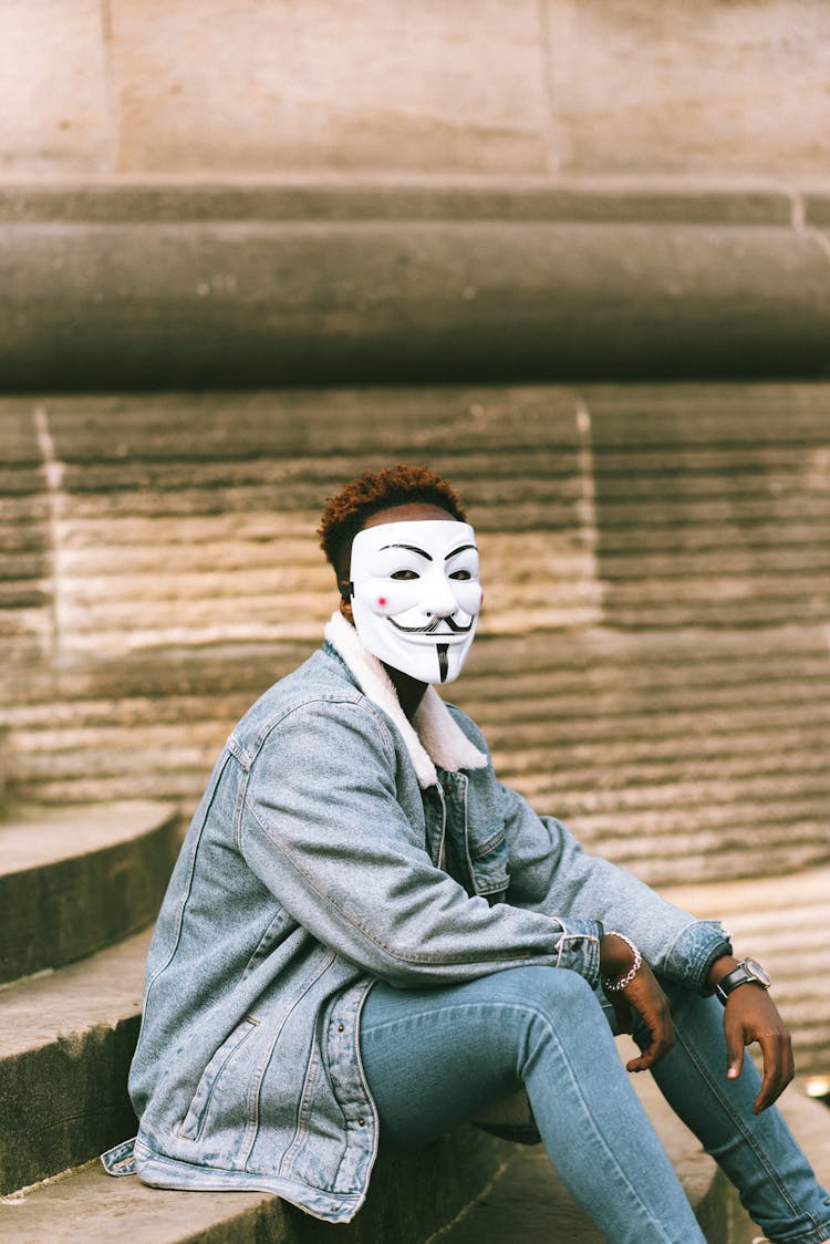 Pensive Black Man In Anonymous Mask Sitting On Street