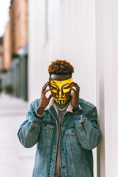 Unrecognizable African American man in stylish denim clothes putting golden Anonymous mask of face as symbol of protest while standing on street in daytime