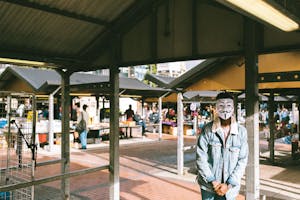 Unrecognizable black man in stylish clothes and Anonymous mask standing in local market in city and looking at camera in sunny day