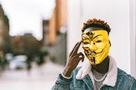 Unrecognizable African American male protester with golden anonymous mask touching temple in town looking at camera