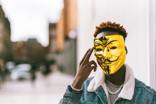 Unrecognizable African American male protester with golden anonymous mask touching temple in town looking at camera