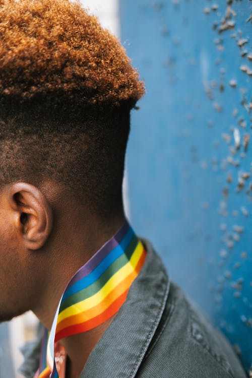 Side view anonymous African American male in denim jacket wearing bright rainbow flag on neck while leaning on grunge blue wall