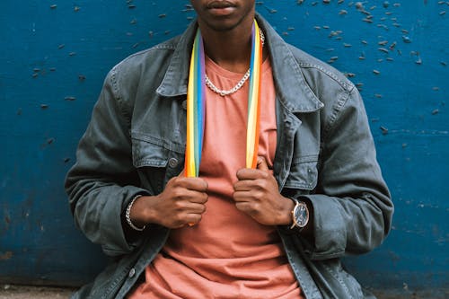 Free Crop emotionless African American male in street style clothes wearing colorful LGBT flag on neck while sitting against shabby blue wall Stock Photo