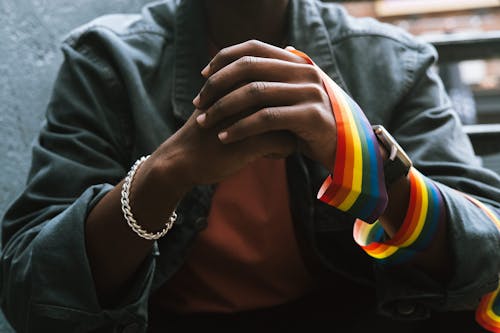 Free Crop anonymous African American homosexual male in casual clothes wearing LGBT flag on arm while sitting on street and clasping hands Stock Photo