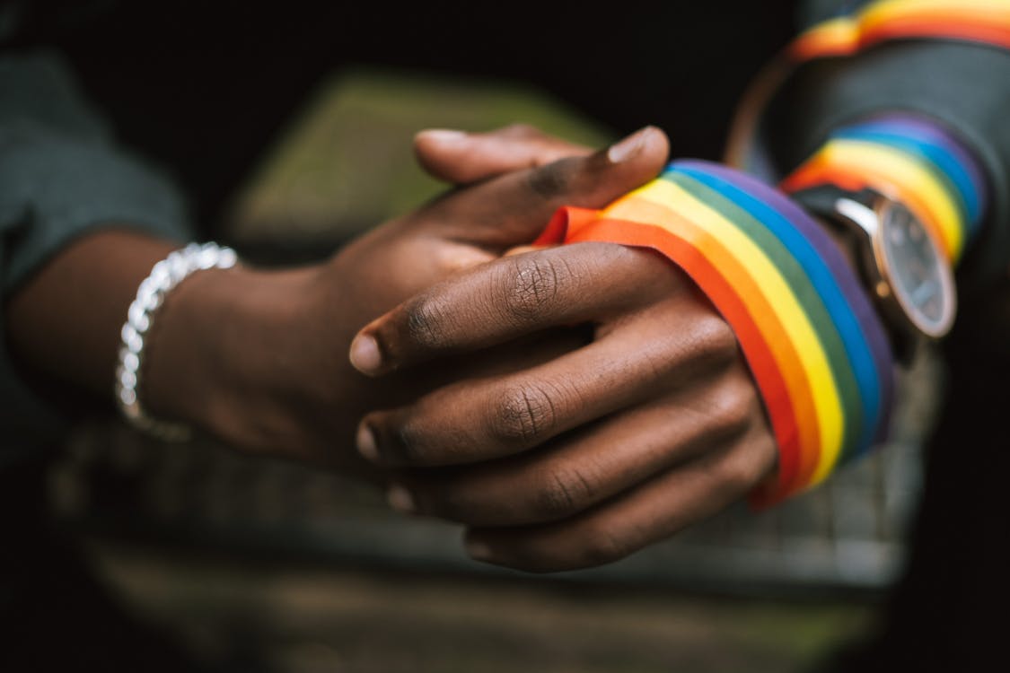 Crop anonymous African American homosexual male wearing colorful rainbow ribbon on arm while sitting on street with hands clasped