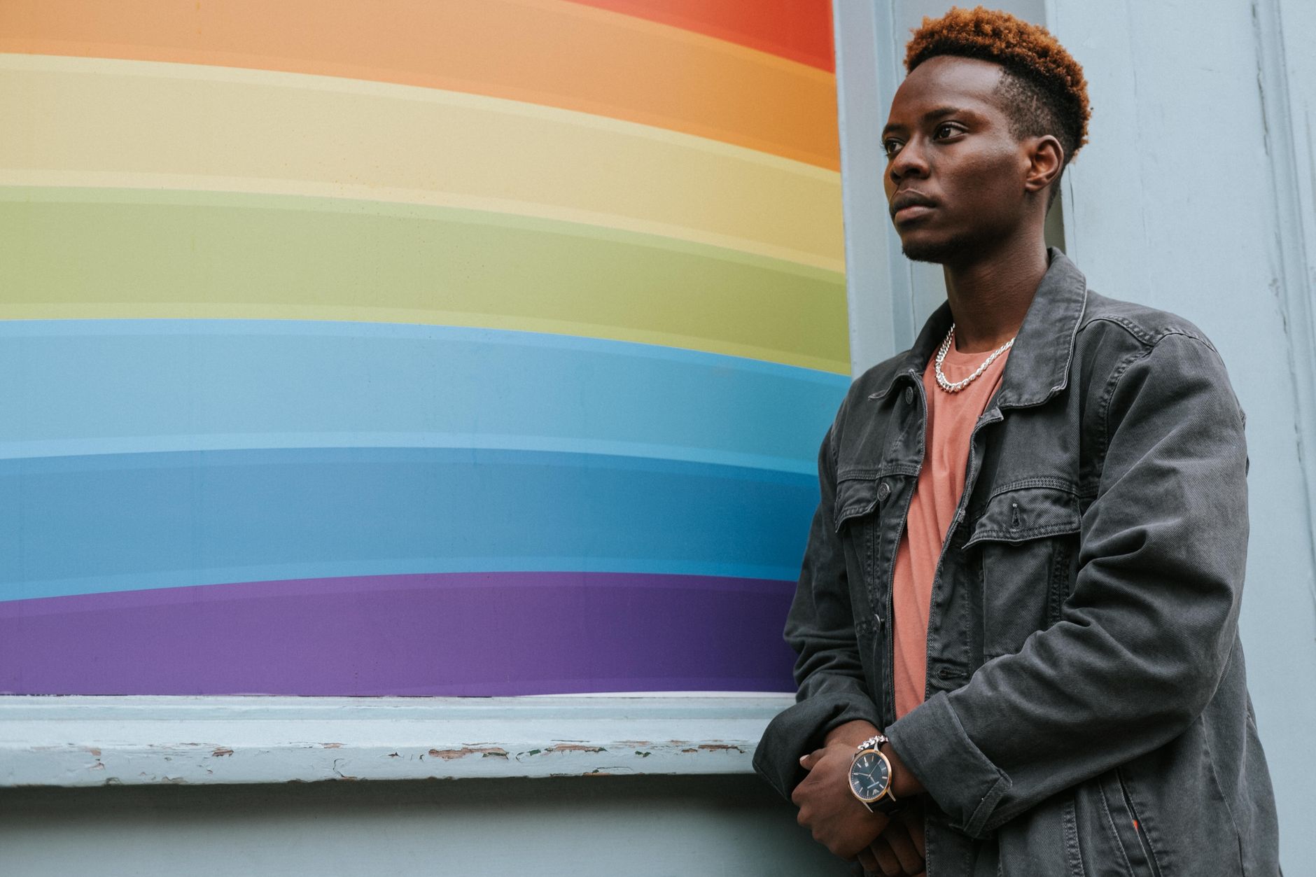 Low angle of pensive young African American guy in trendy outfit standing near wall with colorful LGBT flag and looking away