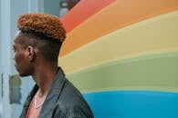 Side view of young black male millennial with trendy haircut in casual outfit standing near wall with LGBT flag and looking away