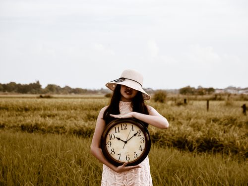 Free A woman in a sundress and hat holds a large wall clock in a rural, grassy field. Stock Photo