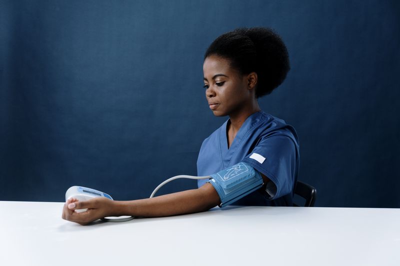 A Black woman in blue scrubs sits at a white table against a dark backdrop with an automatic blood-pressure cuff wrapped around her upper arm, looking calmly at the reading.