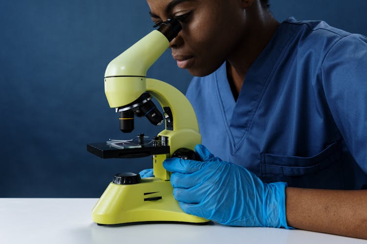 A Woman In Scrubs Looking Through A Microscope