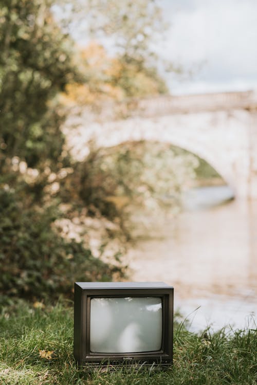Small black vintage television placed on grassy coast near river against lush tree and stone bridge on blurred background in nature