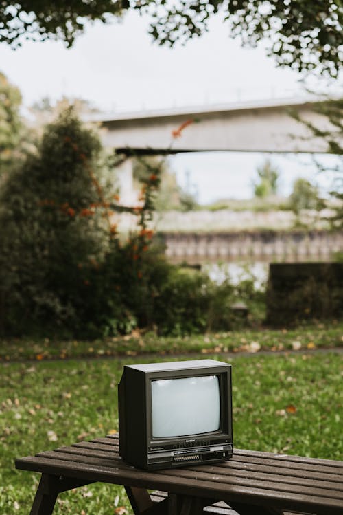 Small black old fashioned television placed on bench in city on street with green plant and bridge on blurred background